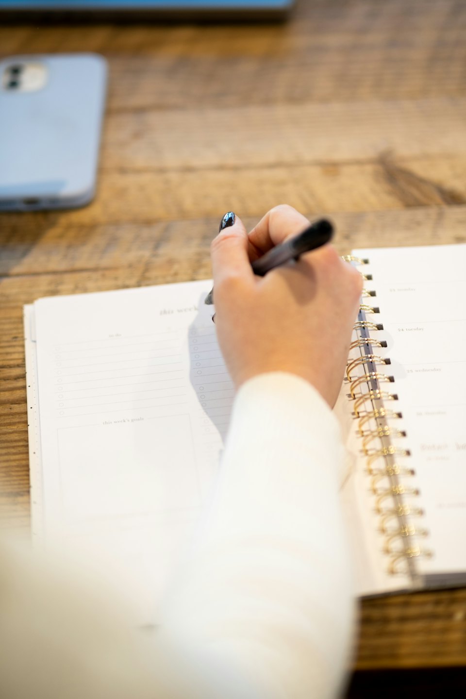 Hand with black nail polish writing in a planner on a wooden desk.